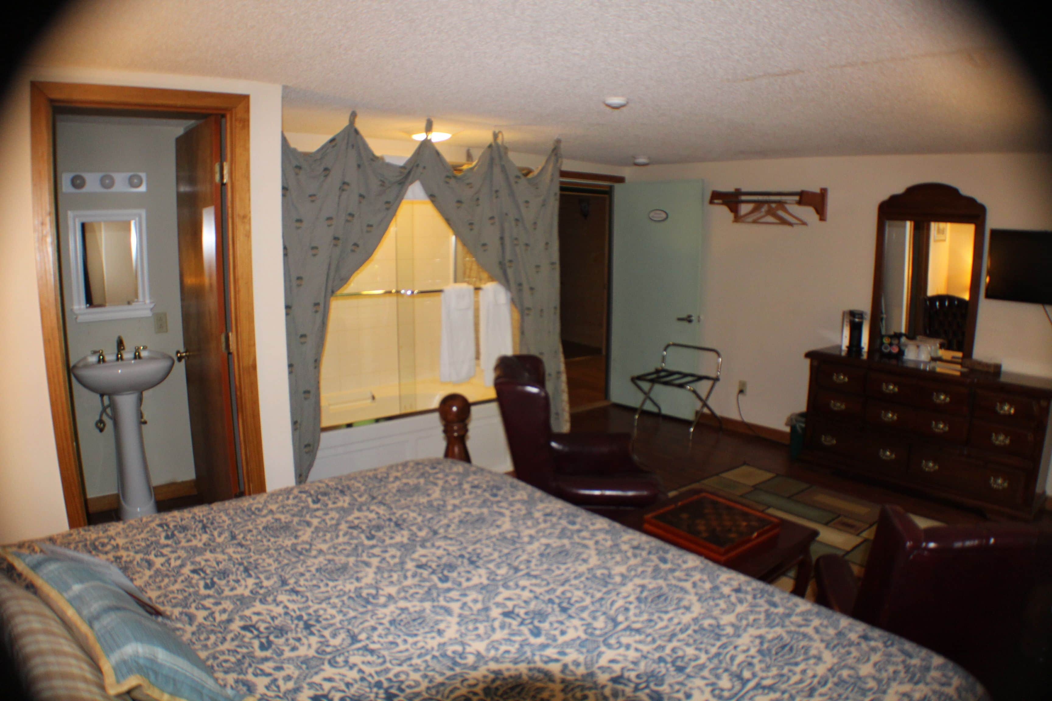 A bedroom with a large bed, a chair, a dresser, a mirror, and a bathroom visible through a window.