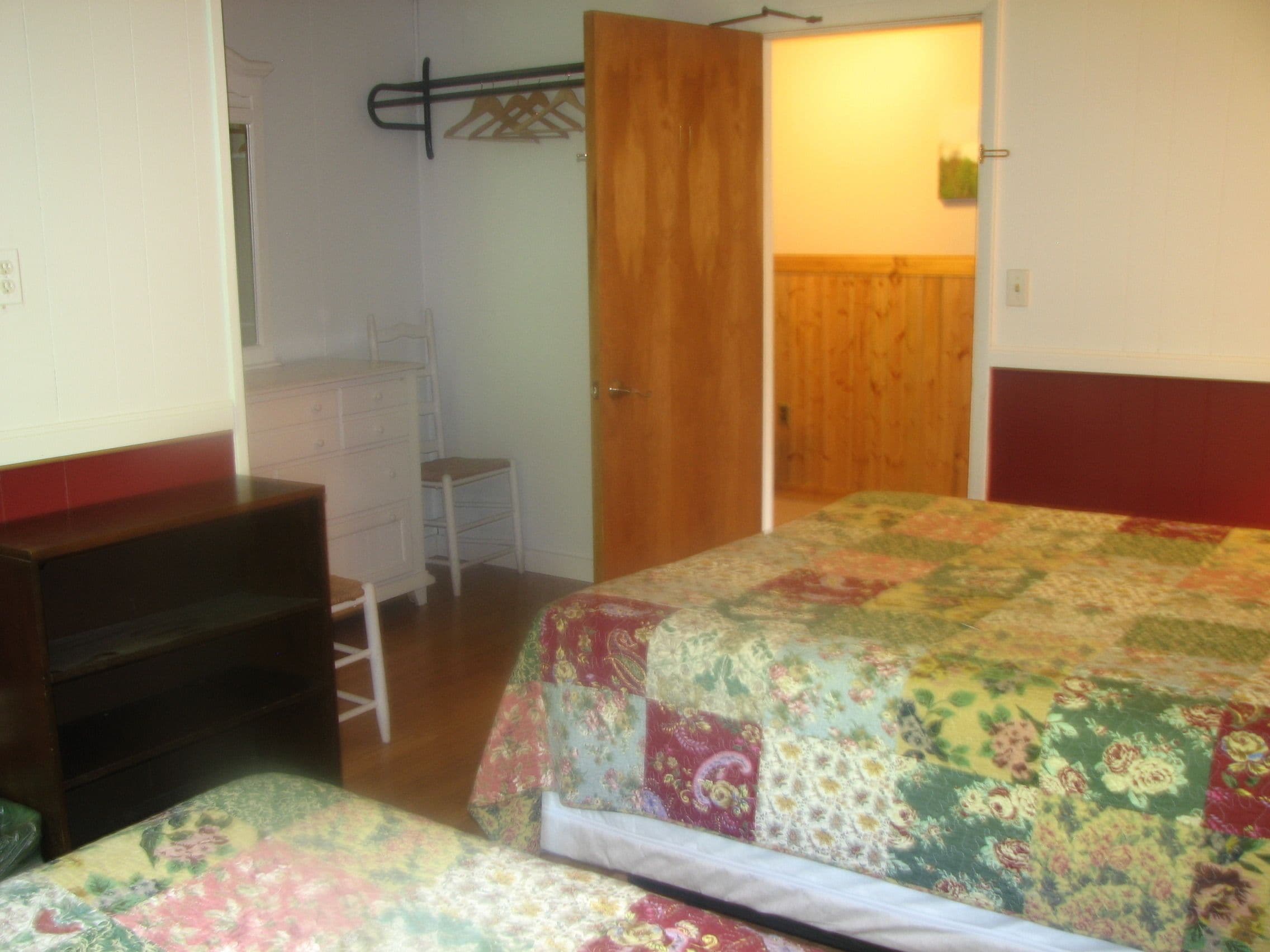 A bedroom with a wooden wardrobe, a bed with a floral quilt, a small white dresser, and a chair.