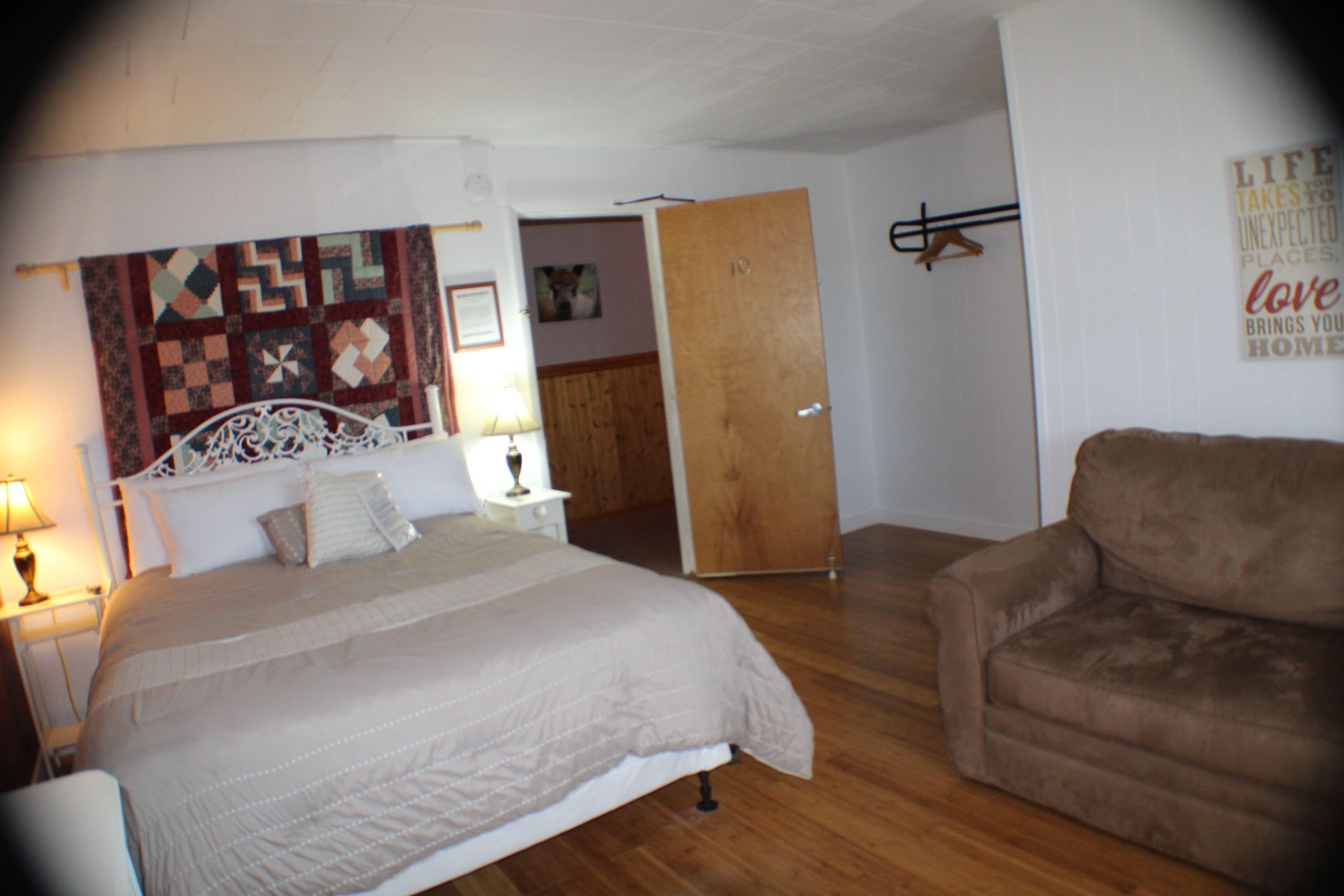 Une chambre à coucher avec un lit, une chaise, un plancher en bois, une porte et un mur orné d’une courtepointe suspendue.