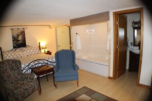 A bedroom with a bed, a chair, a small table, a bathroom with a bathtub, and a shower.
