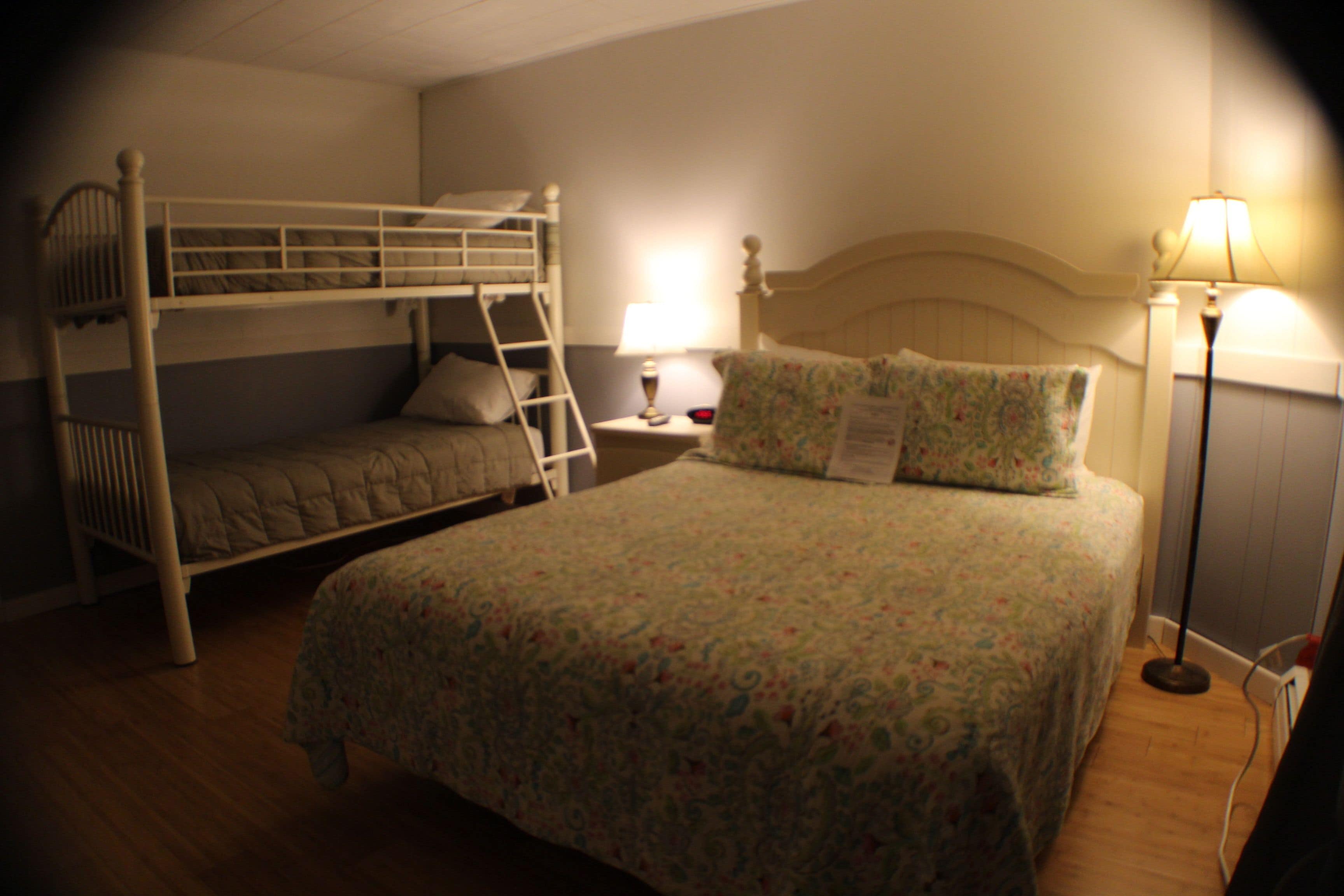 A room with a bunk bed, a single bed, a bedside table with a lamp, and a floor lamp.