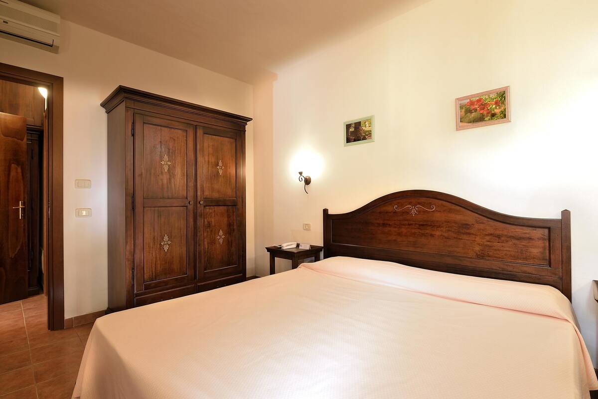 Un dormitori amb un armari de fusta gran, una tauleta de nit amb una làmpada i un llit amb capçalera.