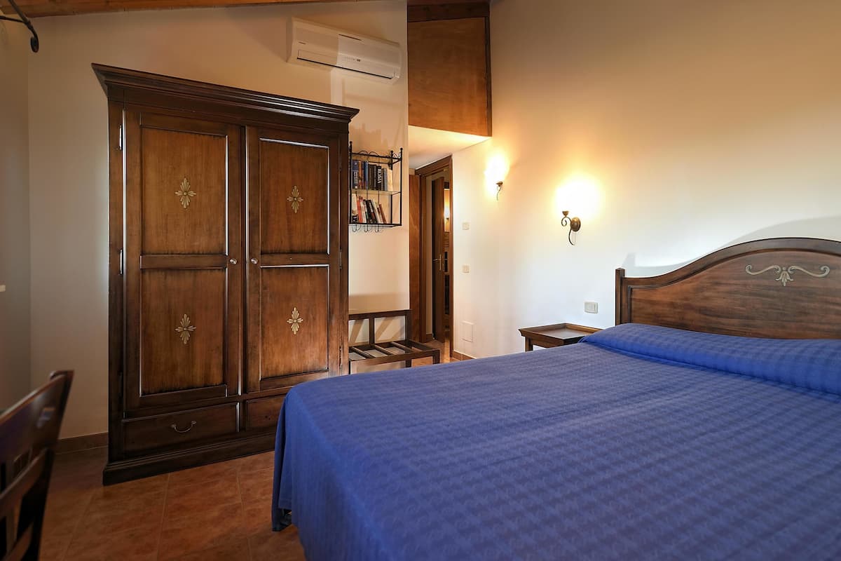 Un dormitori amb un armari de fusta gran, un llit amb edredó blau, una cadira i una prestatgeria penjada a la paret amb llibres.