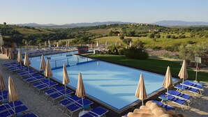 Un espace piscine agrémenté de chaises longues bleues et de parasols, entouré d’un paysage pittoresque avec des collines et un ciel dégagé.