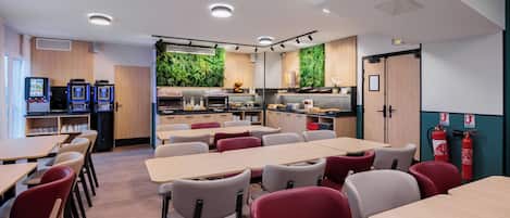 Un espace repas moderne avec plusieurs tables et chaises, un coin cuisine agrémenté d’un mur végétal et des distributeurs automatiques.