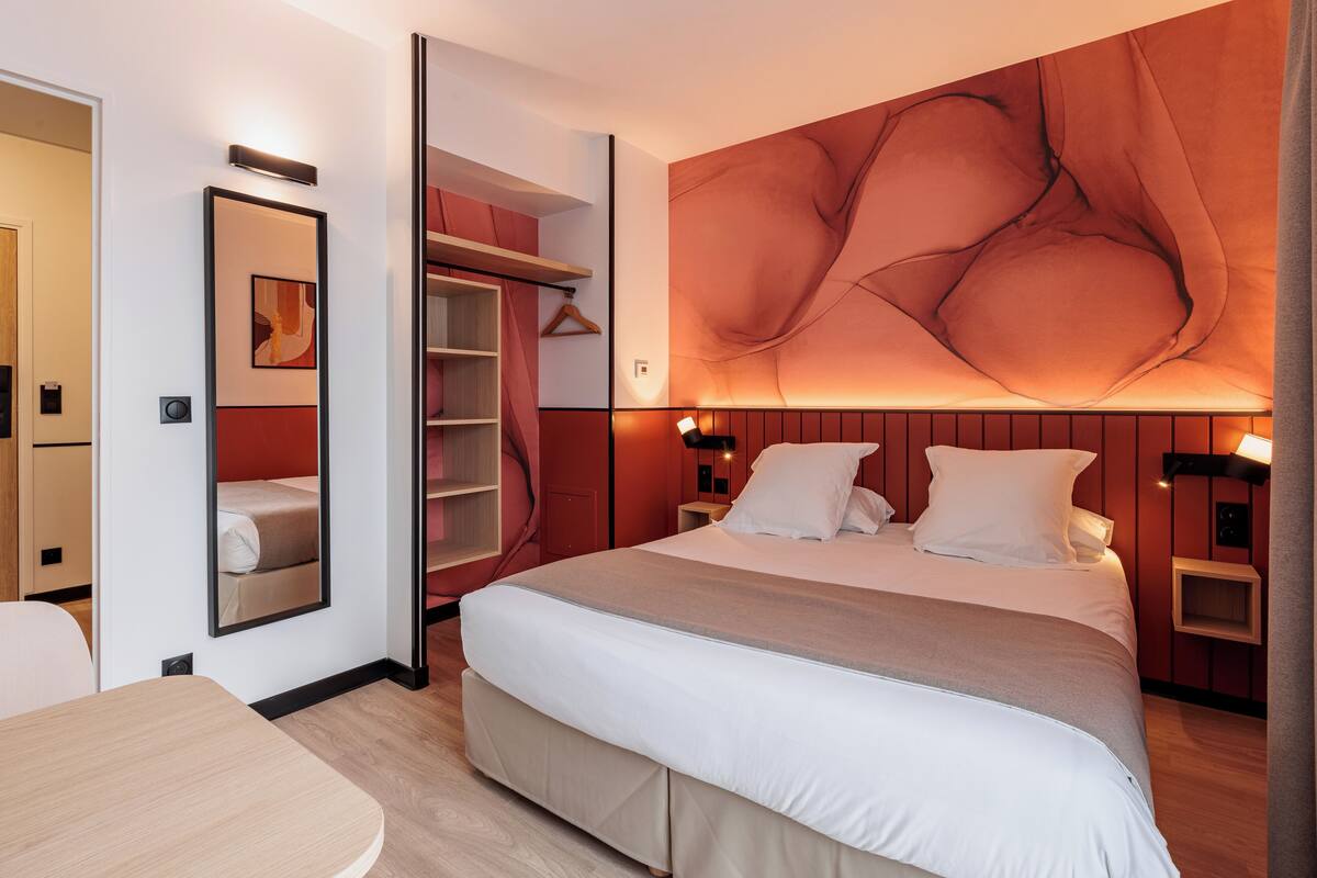 A modern hotel room with a large bed, a bedside table, a wardrobe, and a mirror.