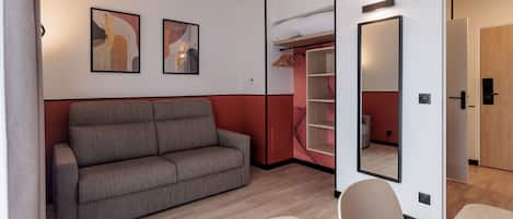 Una moderna camera d'albergo con un divano grigio, pavimento in legno, uno specchio e un armadio pieno di vestiti.