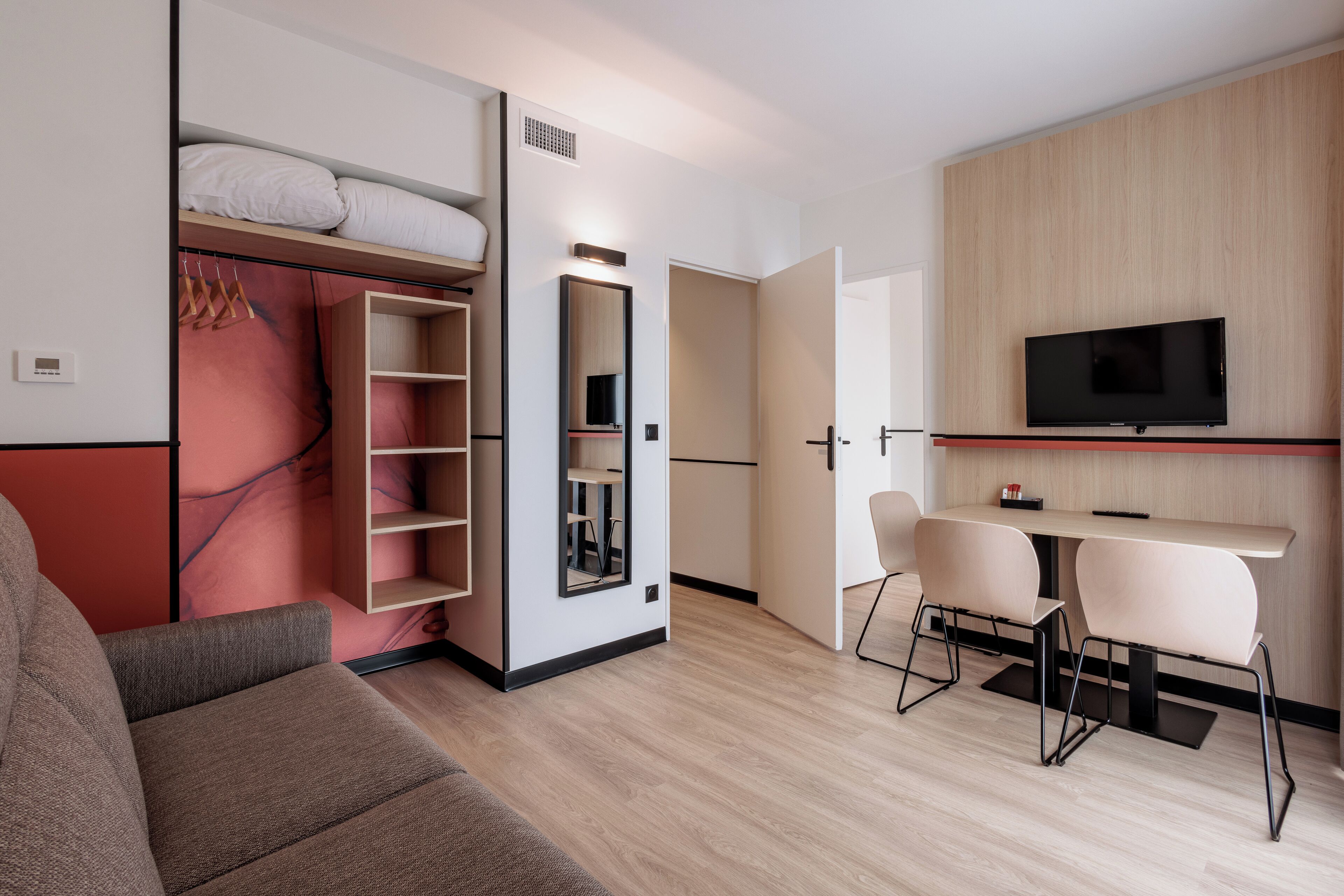 A modern hotel room with a sofa, a TV mounted on the wall, a desk with chairs, and a wardrobe with a mirror.