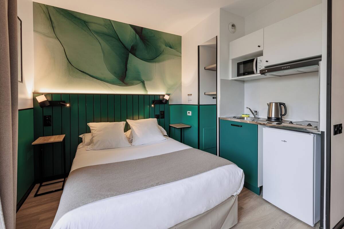 A modern hotel room with a bed, a small kitchenette, and a green accent wall.
