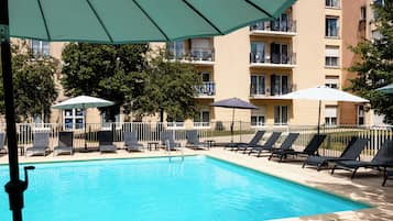 A swimming pool with lounge chairs and umbrellas, surrounded by a fence and a building with balconies.