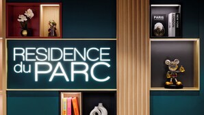 En væg med et neon-skilt, der viser "RESIDENCE du PARC", samt reoler med bøger og dekorative genstande.