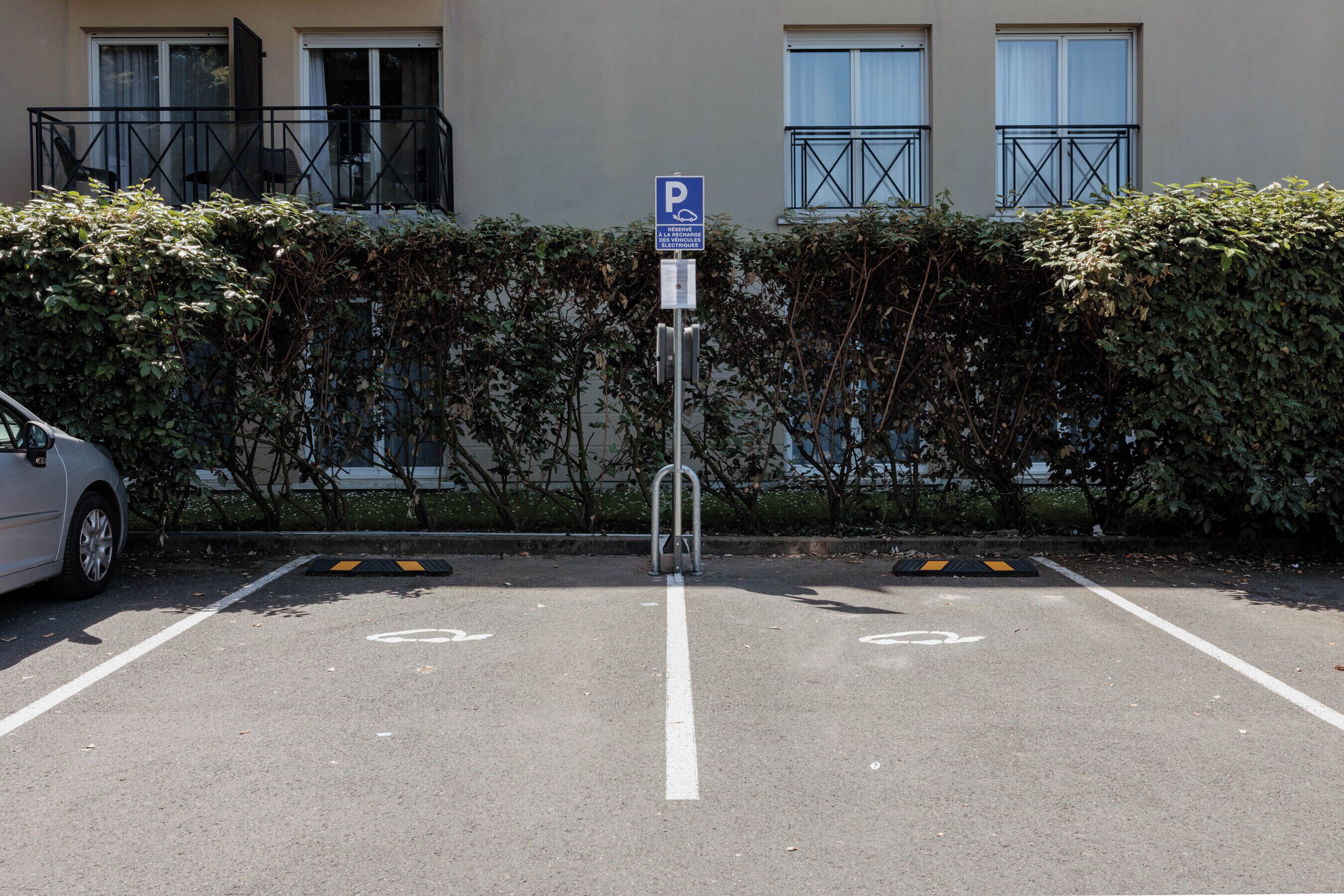 A parking area with a sign indicating a parking spot for disabled individuals, a car parked to the left, and a building with balconies in the background.