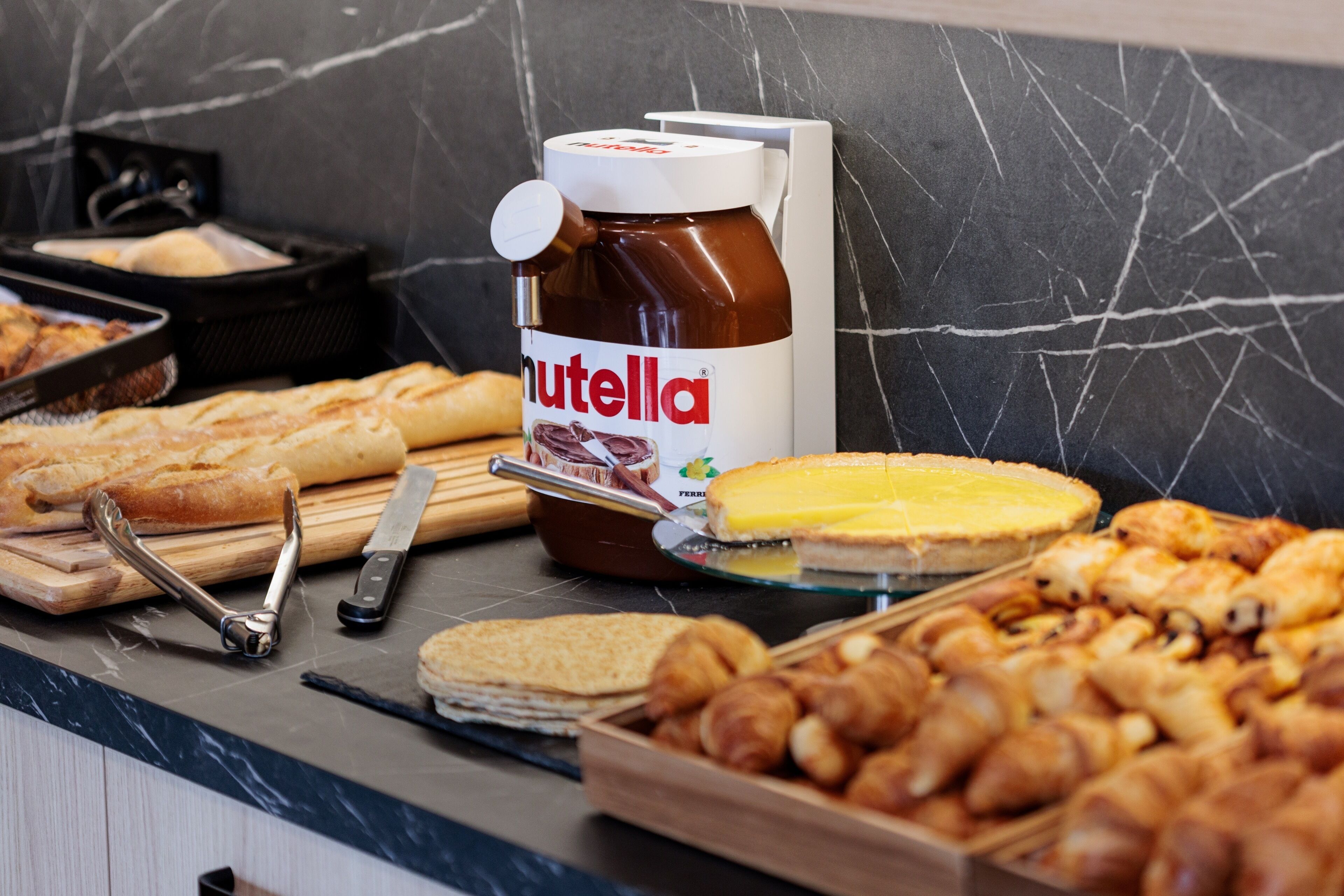 Un buffet proposant diverses pâtisseries, un pot de Nutella et des ustensiles disposés sur un plan de travail en marbre.