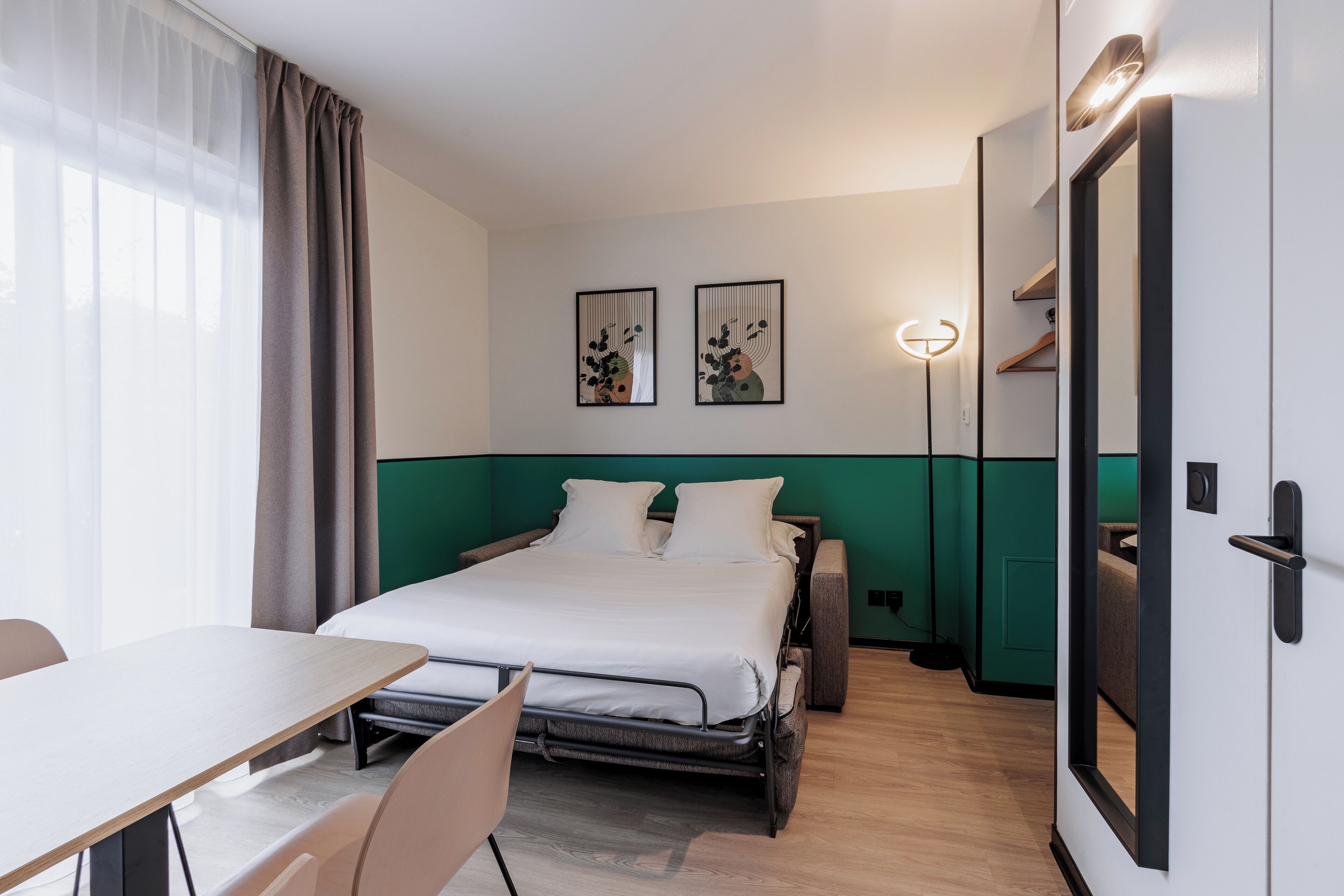 A modern hotel room with a bed, a desk, and a chair. There are two framed artworks on the wall, a mirror, and a door with a handle.