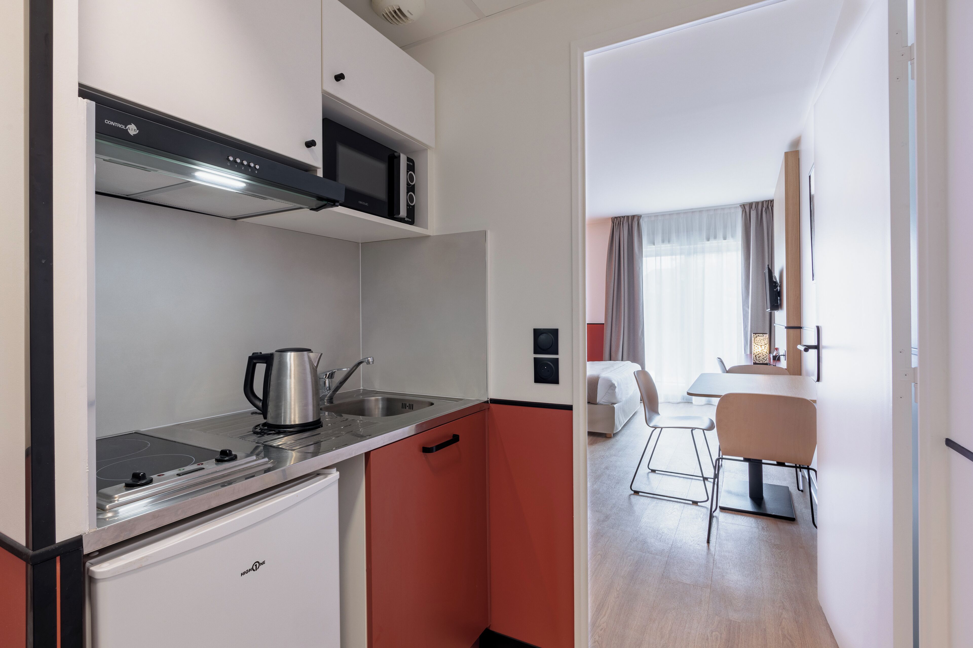 Una cocina compacta con microondas, tetera y un refrigerador pequeño. Se ve una zona de comedor con una mesa y sillas al fondo.