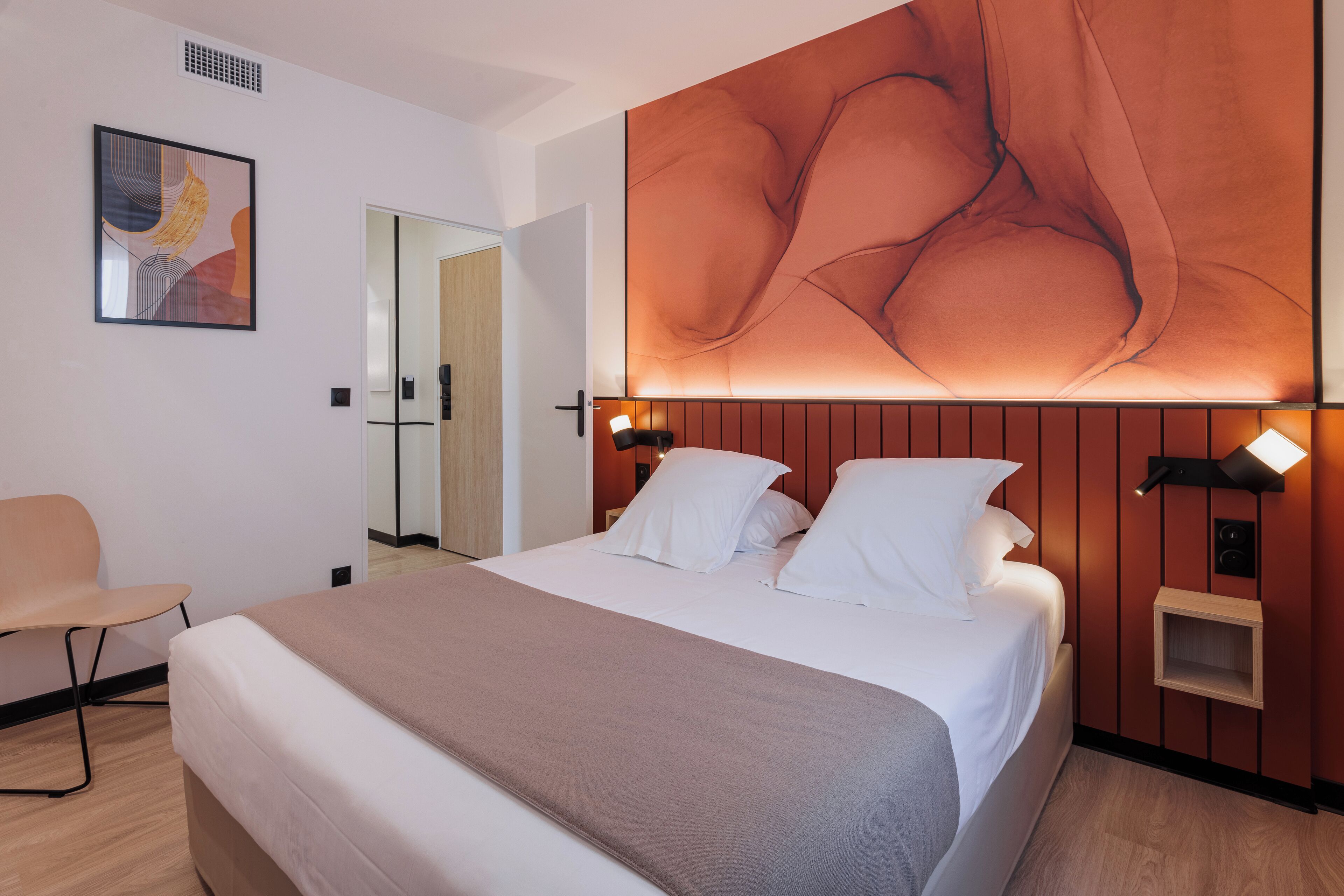 A modern hotel room with a large bed, a bedside table, a chair, and a wall-mounted artwork.