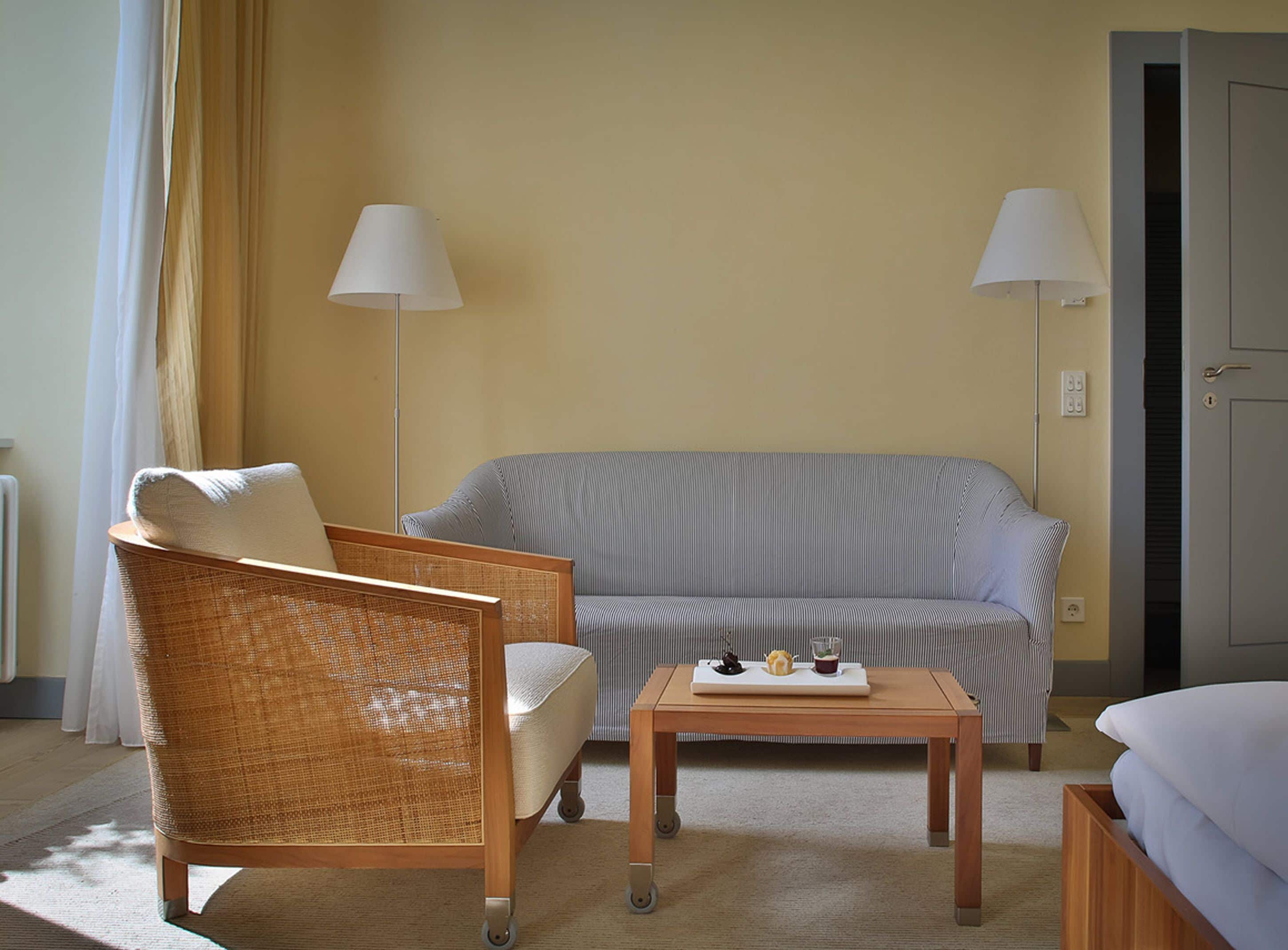 A room with a wicker chair, a striped sofa, a small table with a tray of items, and two floor lamps.