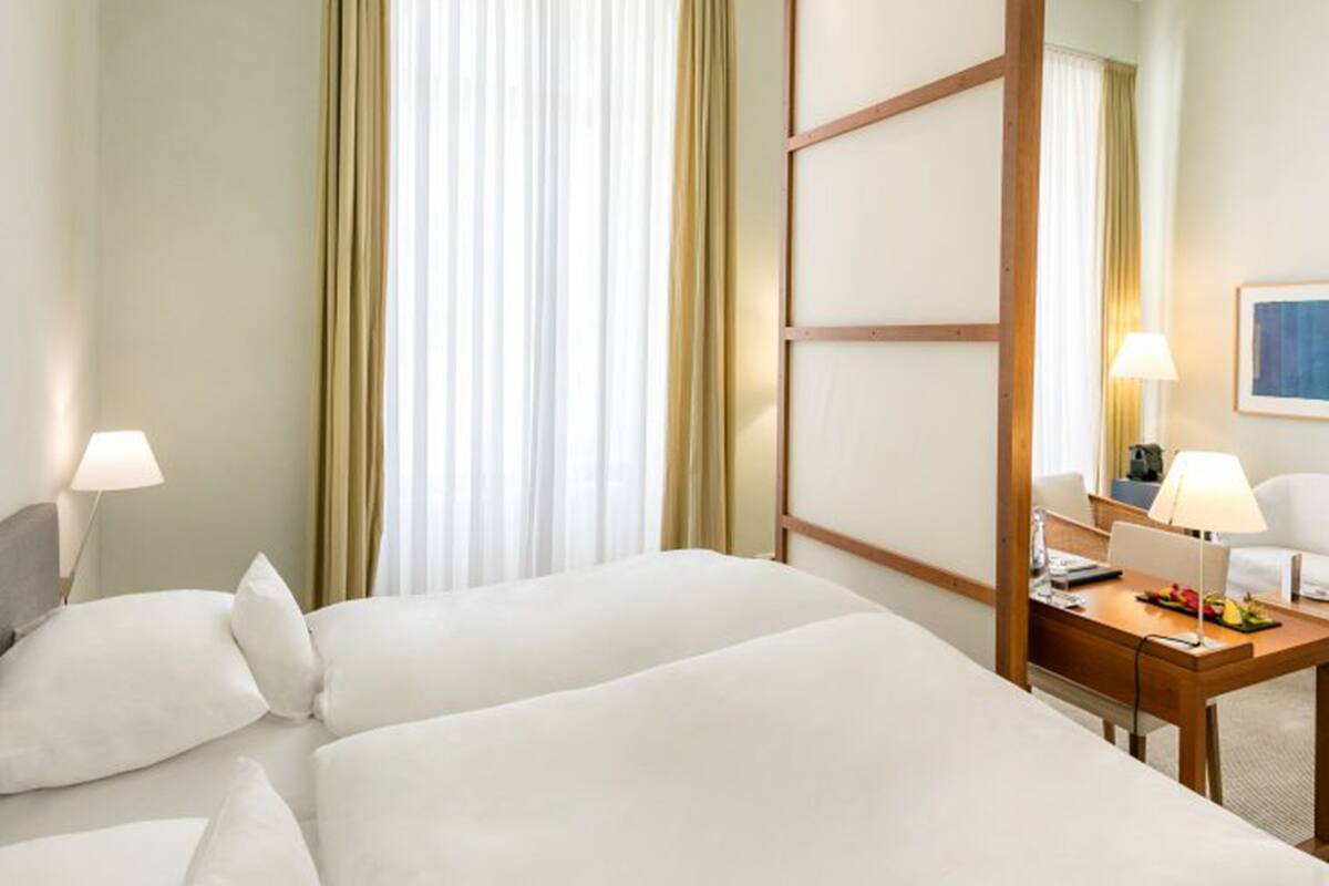 Habitación de hotel con dos camas, un escritorio con lámpara y un amplio ventanal con cortinas.