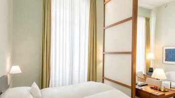 Ein Hotelzimmer mit zwei Betten, einem Schreibtisch mit Lampe und einem großen Fenster mit Vorhängen.