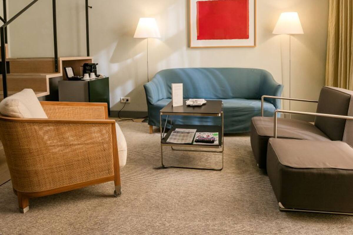 Habitación de hotel moderna con un sofá azul, un sillón de mimbre, una mesita de centro y una lámpara de pie.