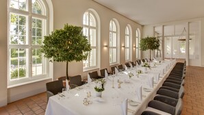 A long dining table set for a formal event, with chairs aligned on both sides, large arched windows, and potted plants.