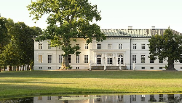 A large, white building with symmetrical architecture, a central entrance, and a surrounding lawn and pond.