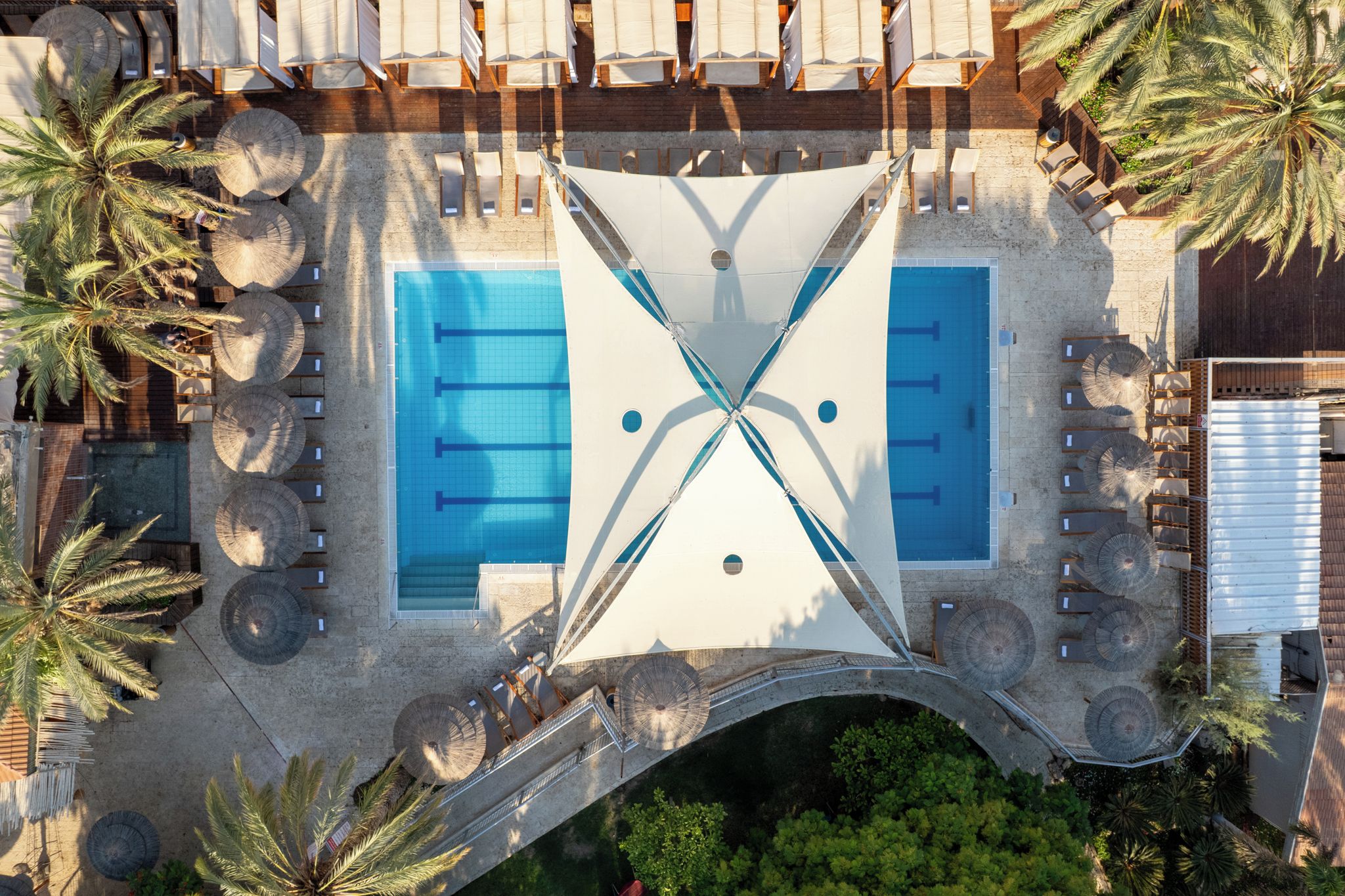 Vista aerea di una zona piscina con due piscine separate, circondata da lettini prendisole e palme.
