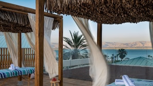 Terraza de madera con dos camas, cada una con ropa de cama y toallas a rayas, con vistas a una playa y montañas.