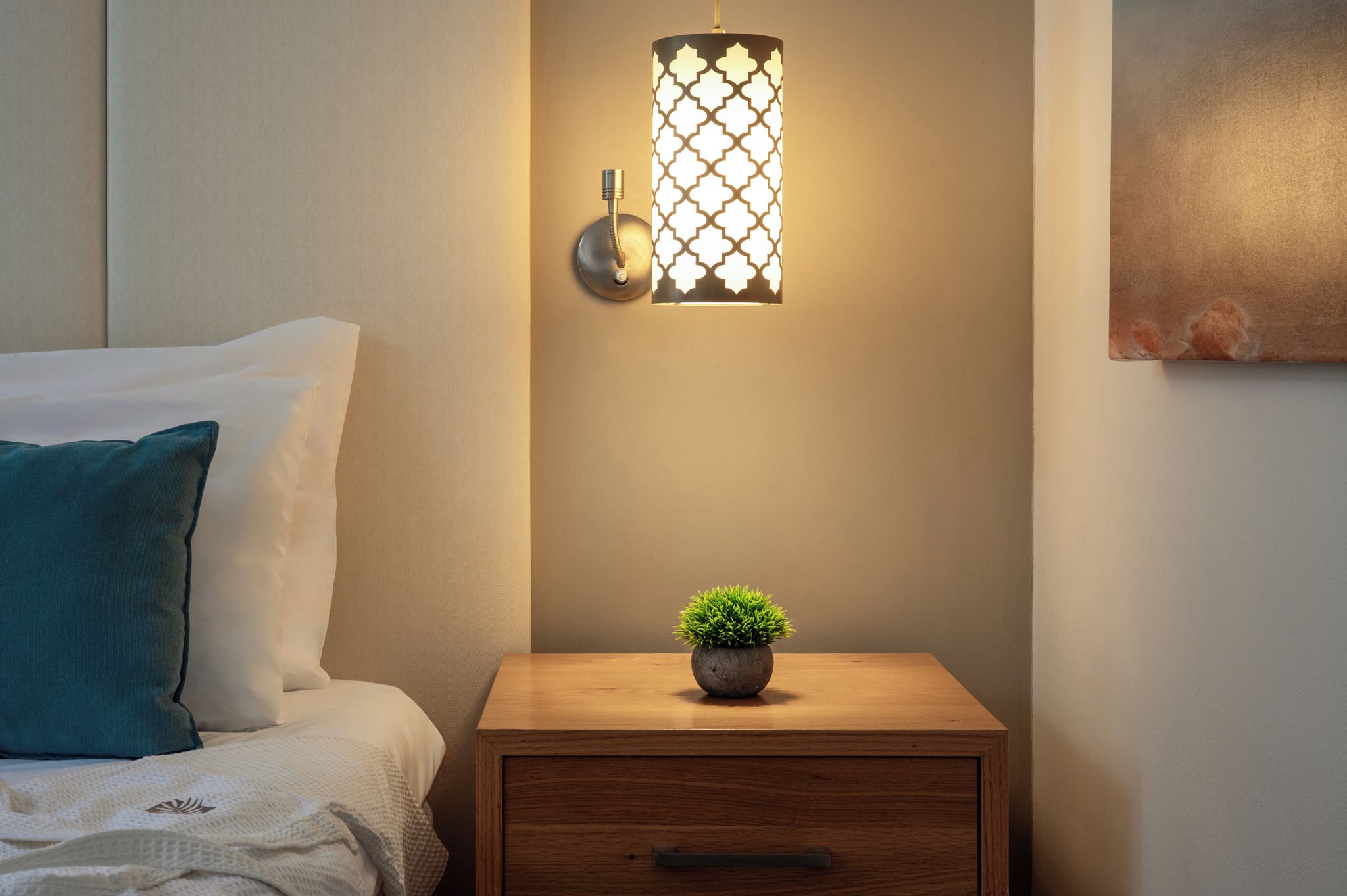 A neatly made bed with a teal pillow, a bedside table with a small potted plant, and a wall-mounted light fixture.