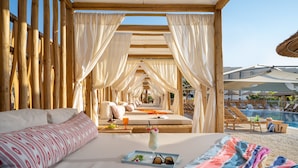 Habitación de hotel con cama, una bandeja con bebida y gafas de sol, y vista a un área de piscina con sillones de descanso.