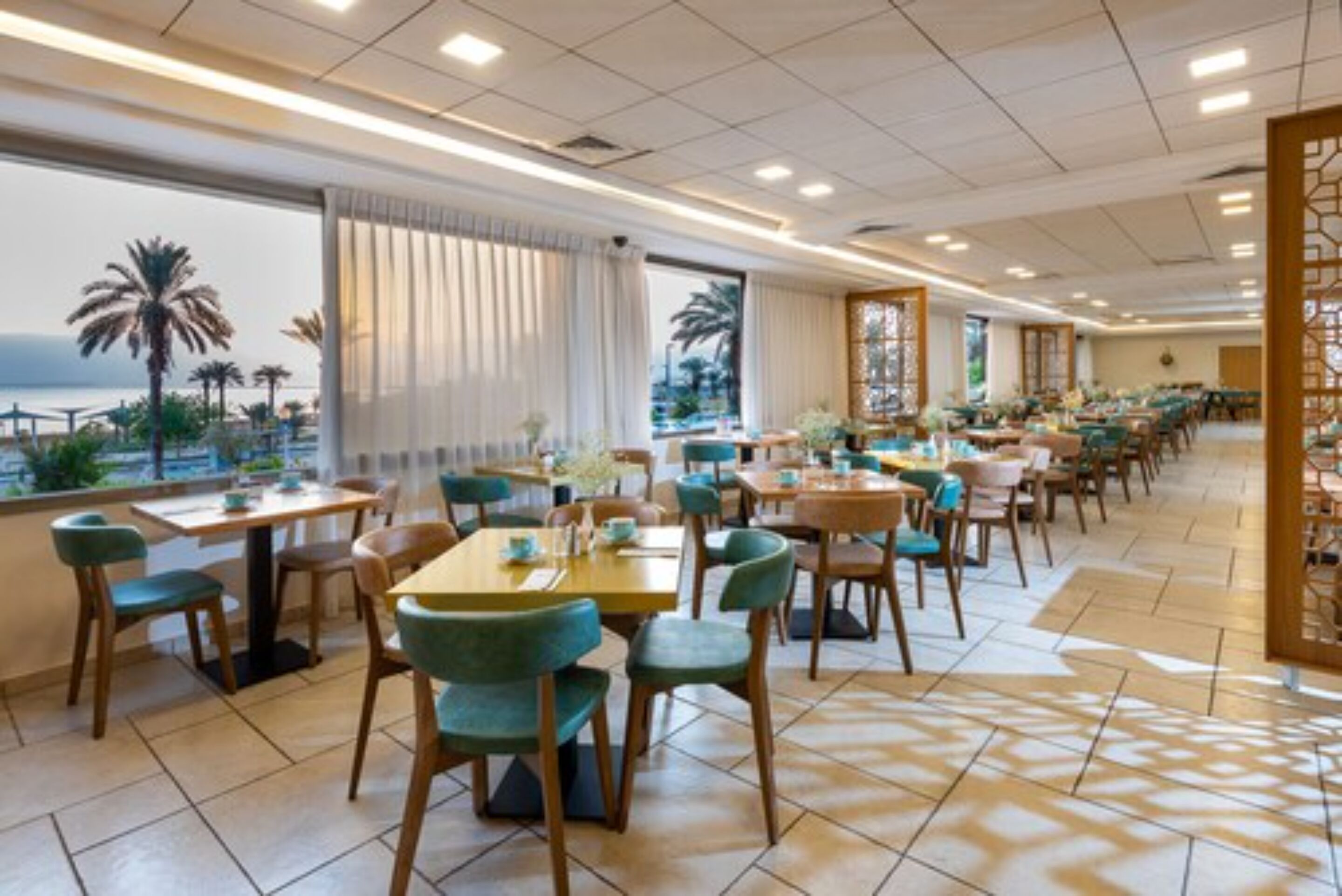 A spacious restaurant with multiple tables and chairs, large windows with curtains, and a view of palm trees and the sea.