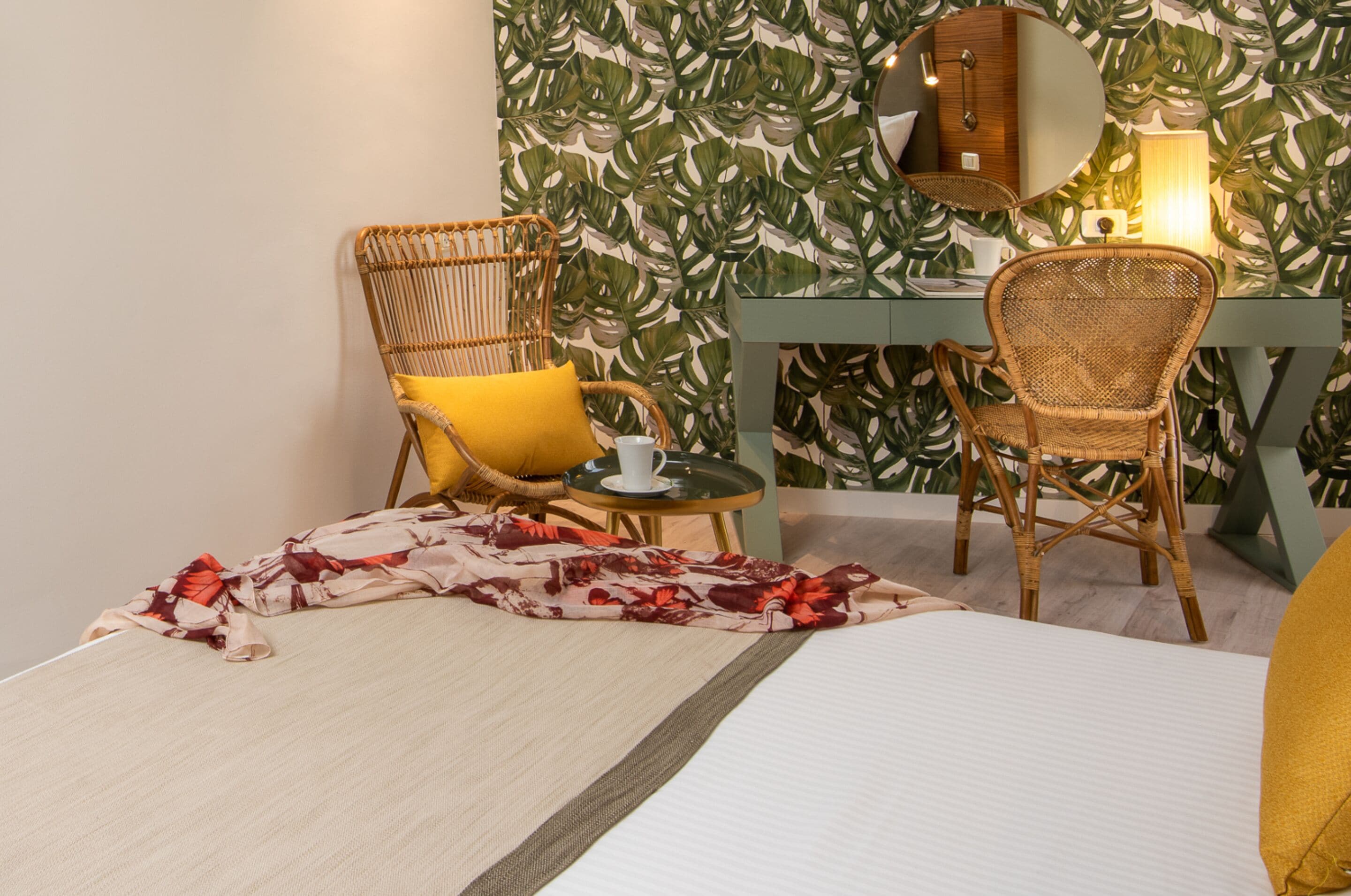 Una habitación con una cama, dos sillones de mimbre, una mesita con una taza y una lámpara, y una pared con un diseño de hojas.