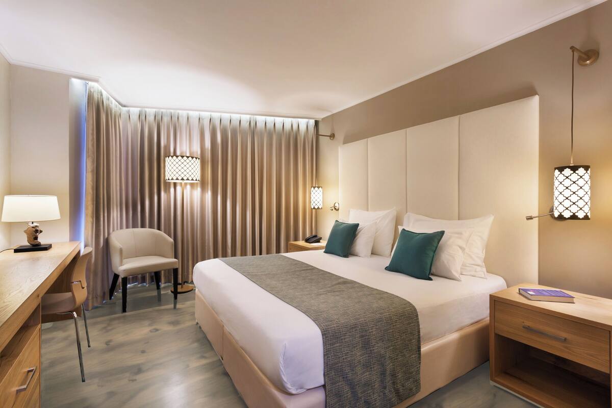 Hotellihuone, jossa on sänky, yöpöydät, työpöytä, tuoli ja ikkuna, jossa on verhot.