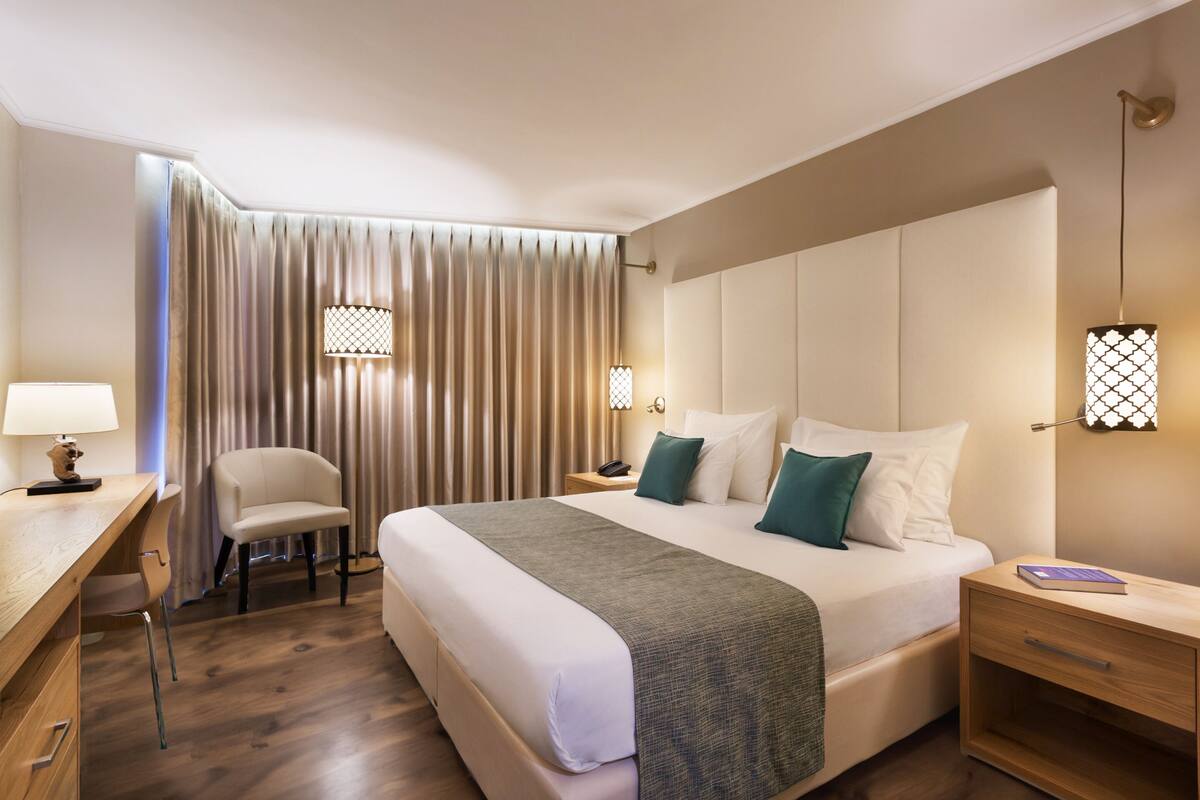 Hotellihuone, jossa on suuri sänky, työpöytä tuolilla, yöpöytä ja seinään kiinnitetty valaisin.