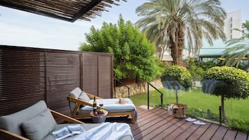 Varanda com sofá, mesa com garrafa e copos, e vista para um jardim com palmeiras e um edifício ao fundo.