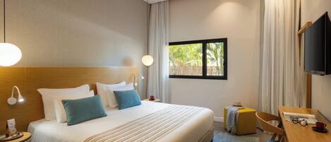 A hotel room with a large bed, bedside lamps, a desk, a chair, a television, and a window with a view of greenery.