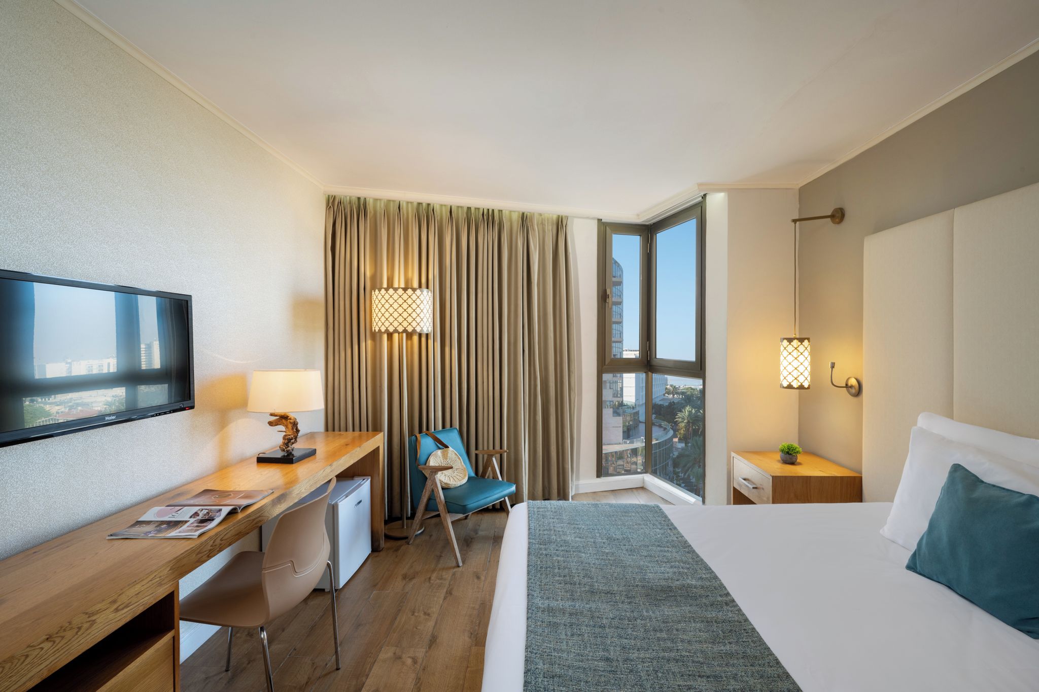 Habitación de hotel con cama, escritorio, silla, televisor, cortinas y ventana con vistas.