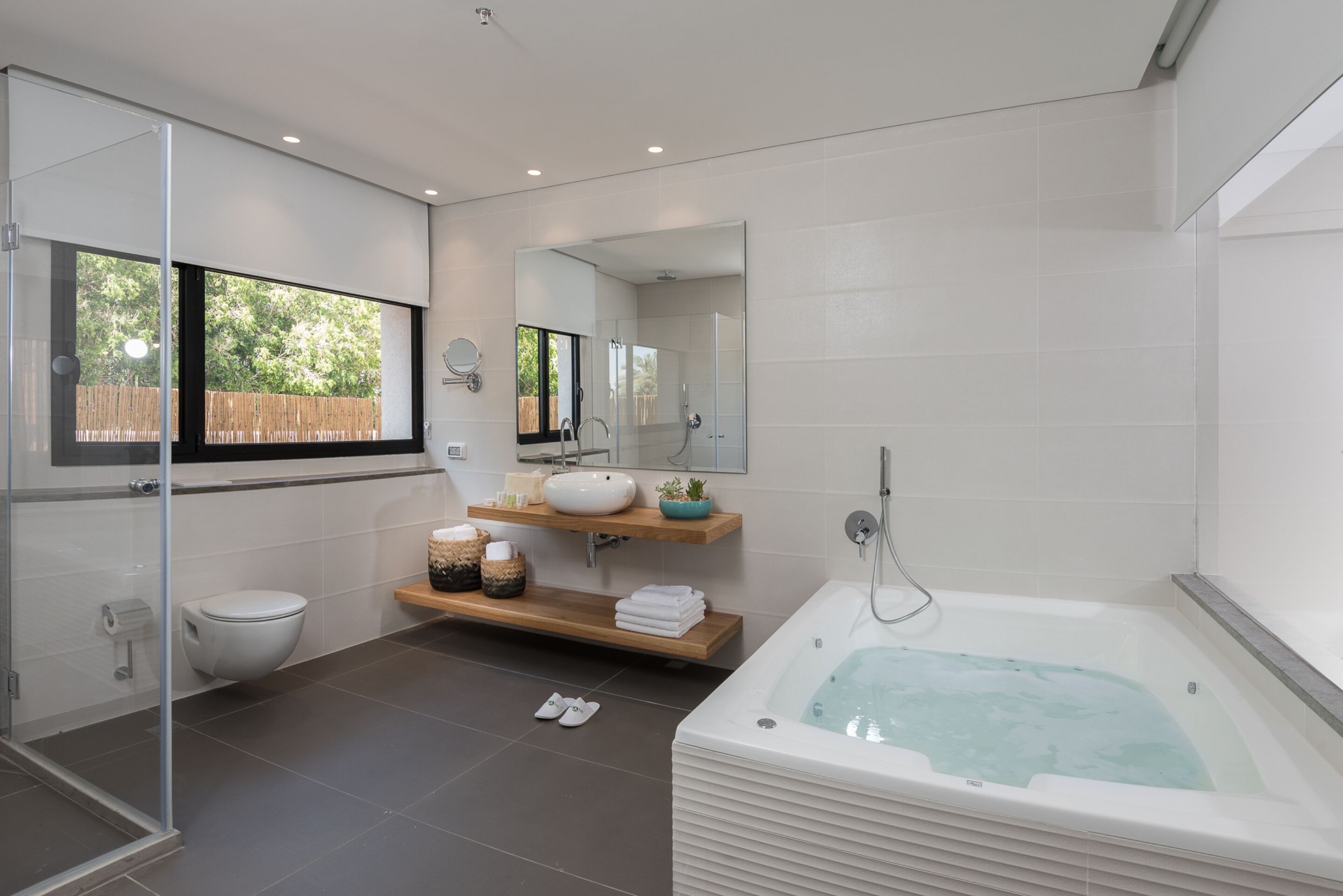 A modern bathroom with a glass shower enclosure, a freestanding bathtub, a large mirror, and a wooden shelf with towels.