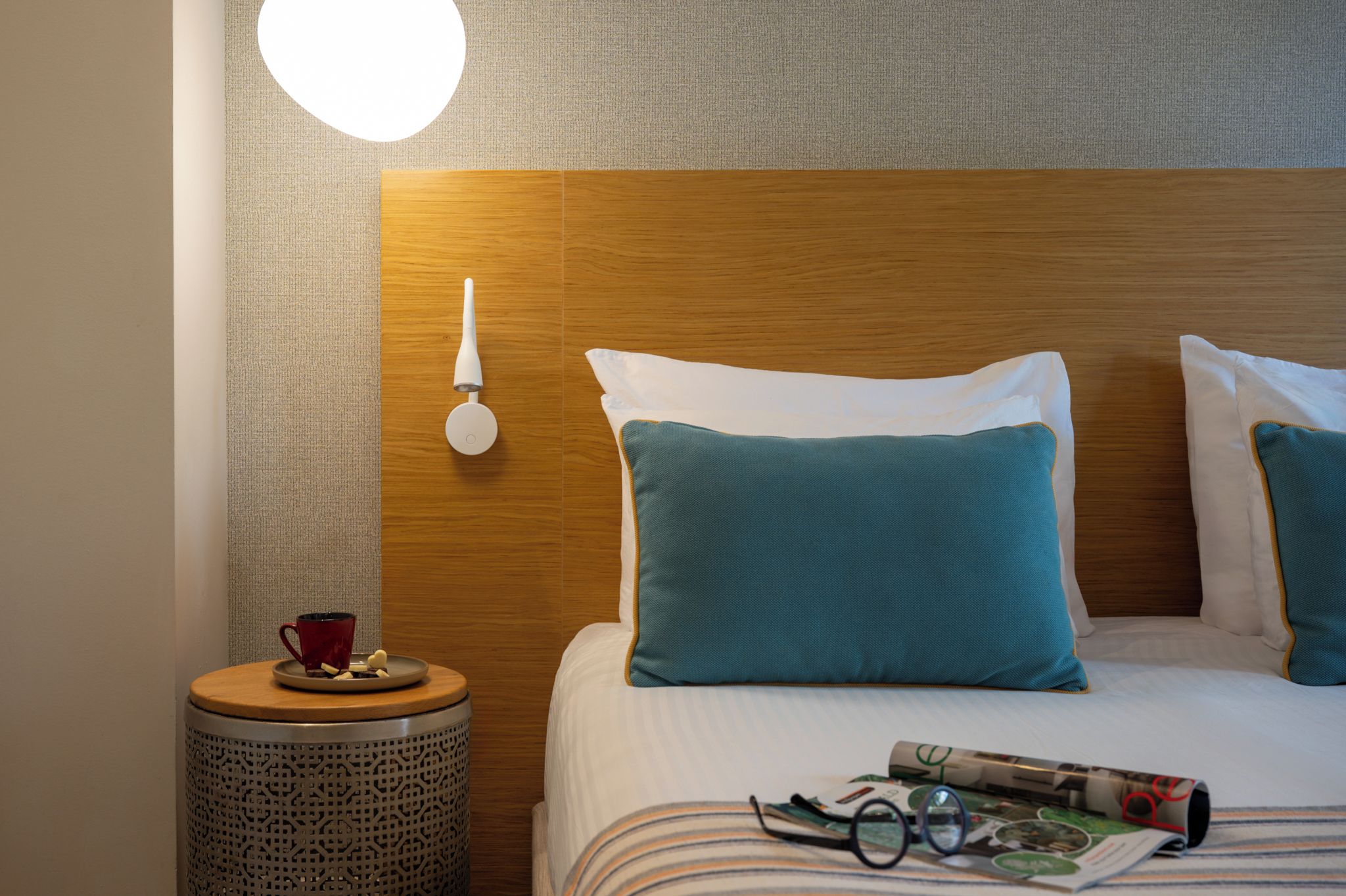 Une chambre d’hôtel avec un lit bien fait, une table de chevet sur laquelle se trouvent une tasse et une assiette, et une lampe fixée au mur.