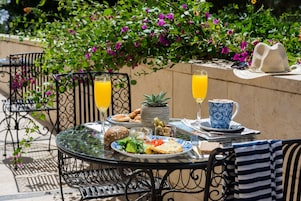Un área de comedor al aire libre con una mesa dispuesta para dos, que presenta una variedad de alimentos y bebidas, rodeada de exuberante follaje y flores.