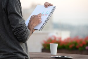 Una persona sosteniendo un cuaderno de espiral con un bolígrafo, una taza blanca y un objeto redondo sobre una mesa de madera.