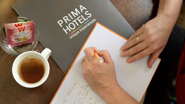 Una persona escribiendo en un cuaderno con un bolígrafo, una taza de té y un folleto de Prima Hotels sobre una mesa de madera.