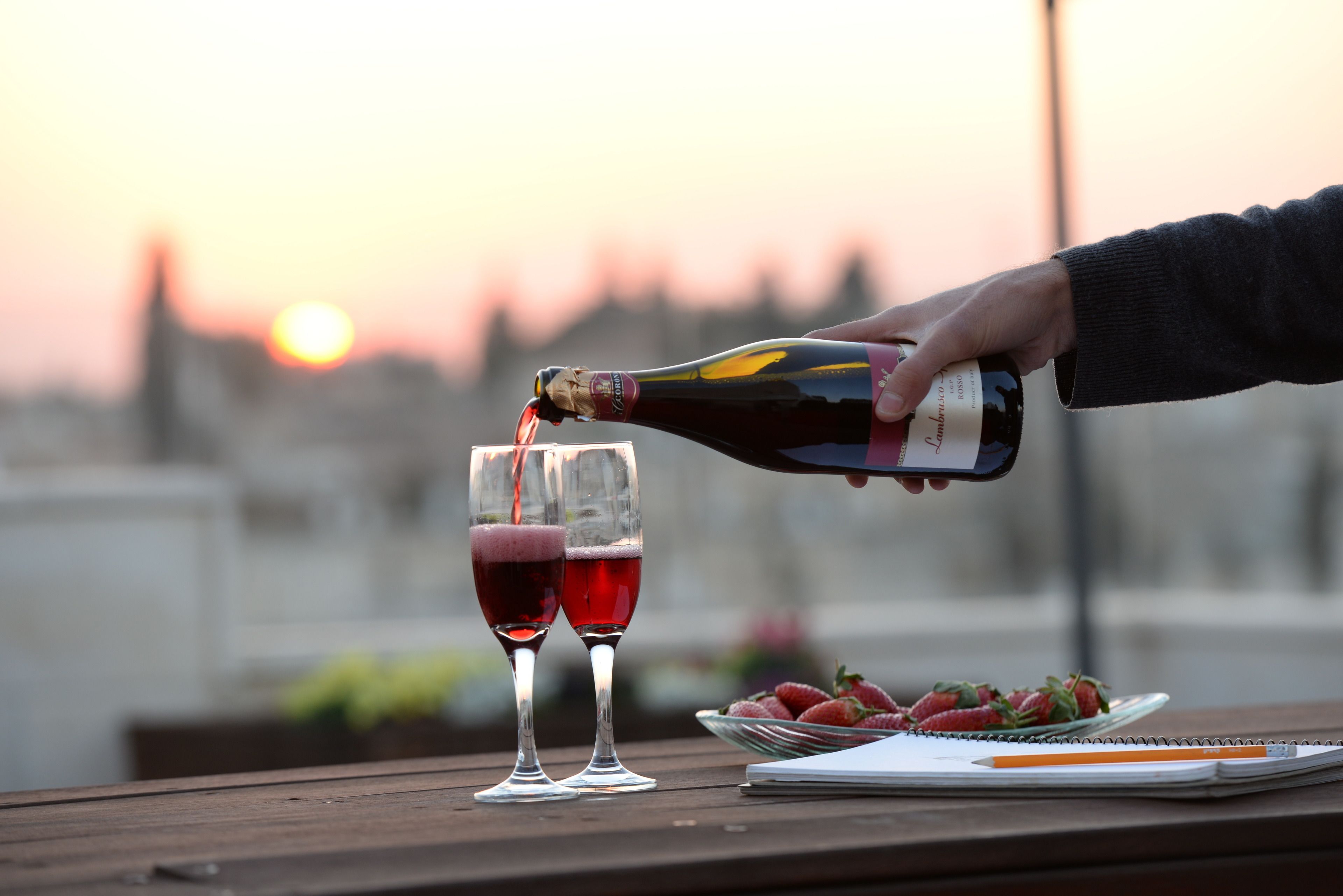 Una botella de champán siendo servida en dos copas sobre una mesa de madera, junto a una libreta y un cuenco de fresas.