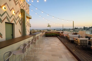 Zona de bar al aire libre con pared de diseño en zigzag, taburetes y luces colgantes.