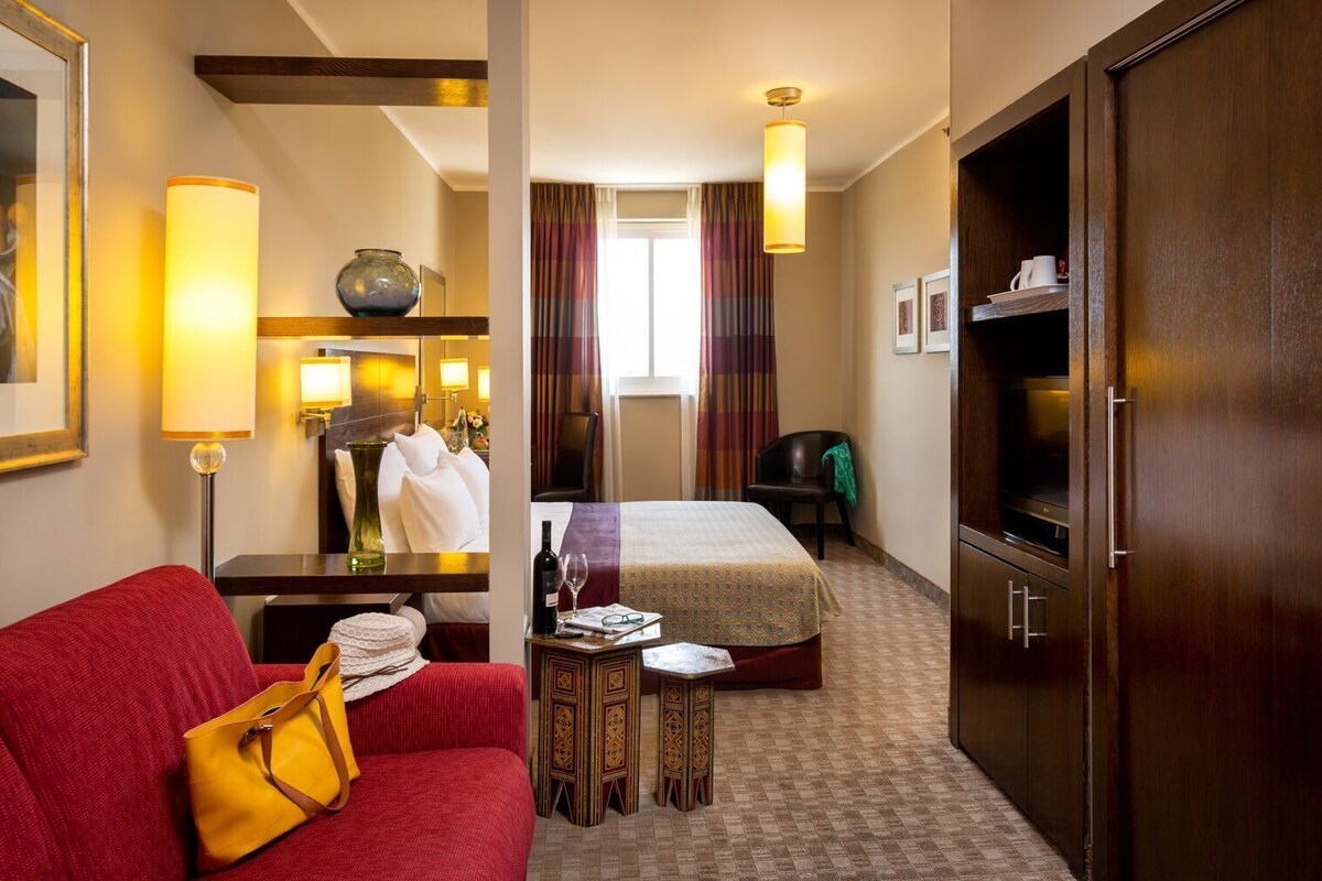 A hotel room with a bed, a red sofa, a small table with a lamp, a TV, and a wardrobe.