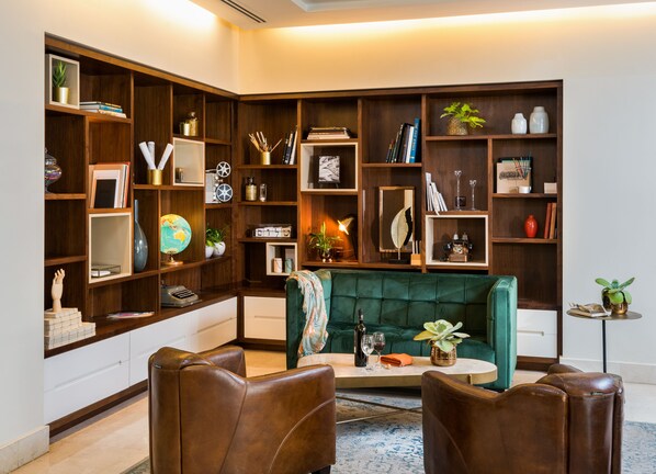 Una sala de estar acogedora con un sofá verde, sillones de cuero y una mesa de centro. Cuenta con estanterías de madera llenas de libros, objetos decorativos y plantas.