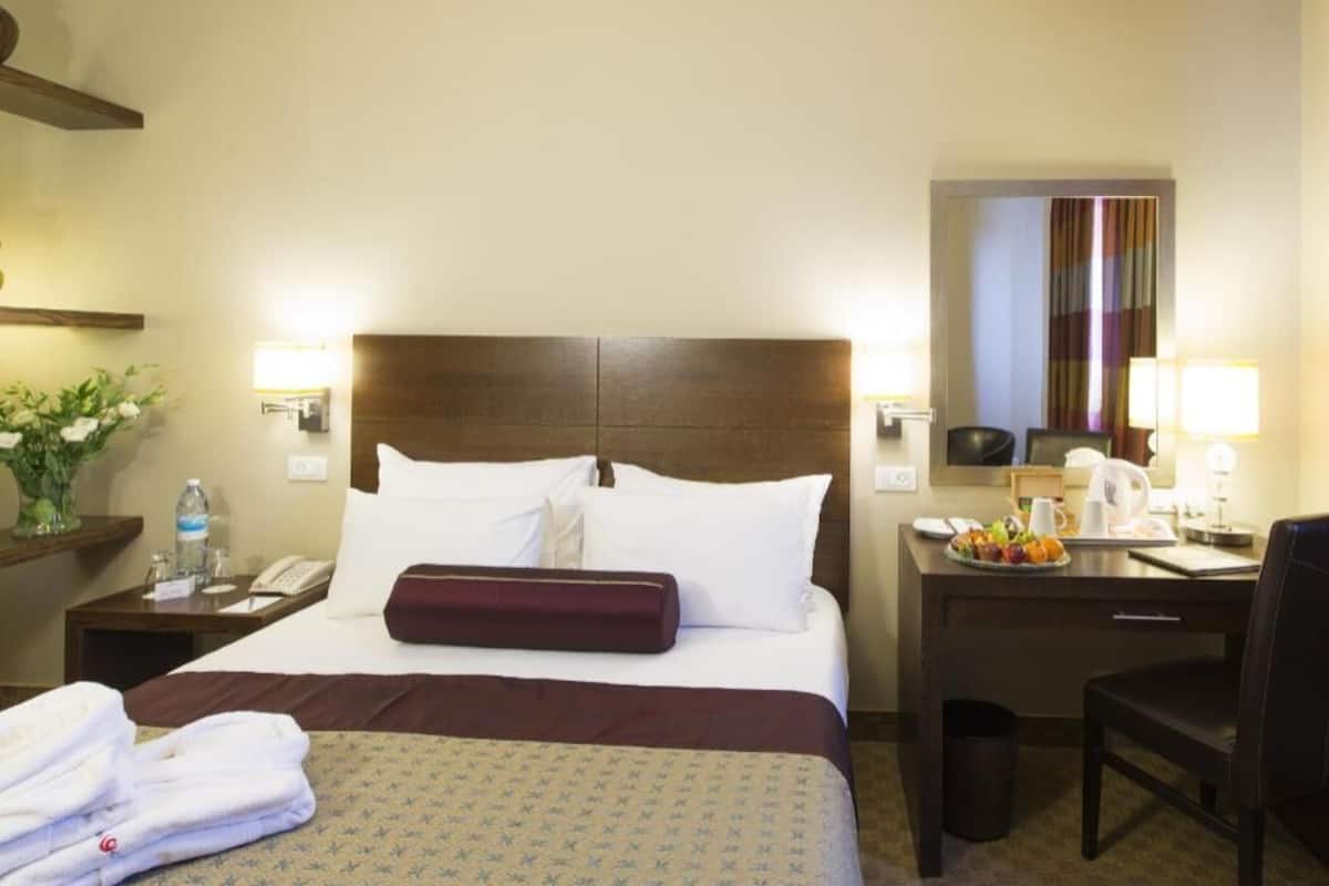 Quarto de hotel com cama, mesas de cabeceira, escrivaninha, cadeira, espelho e ventilador de teto.