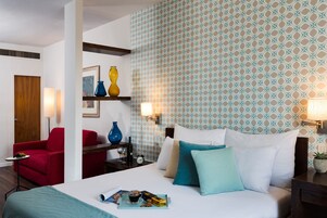 Una habitación de hotel con una cama, un sillón rojo, una mesita de madera, un reloj de pared y un papel tapiz estampado.