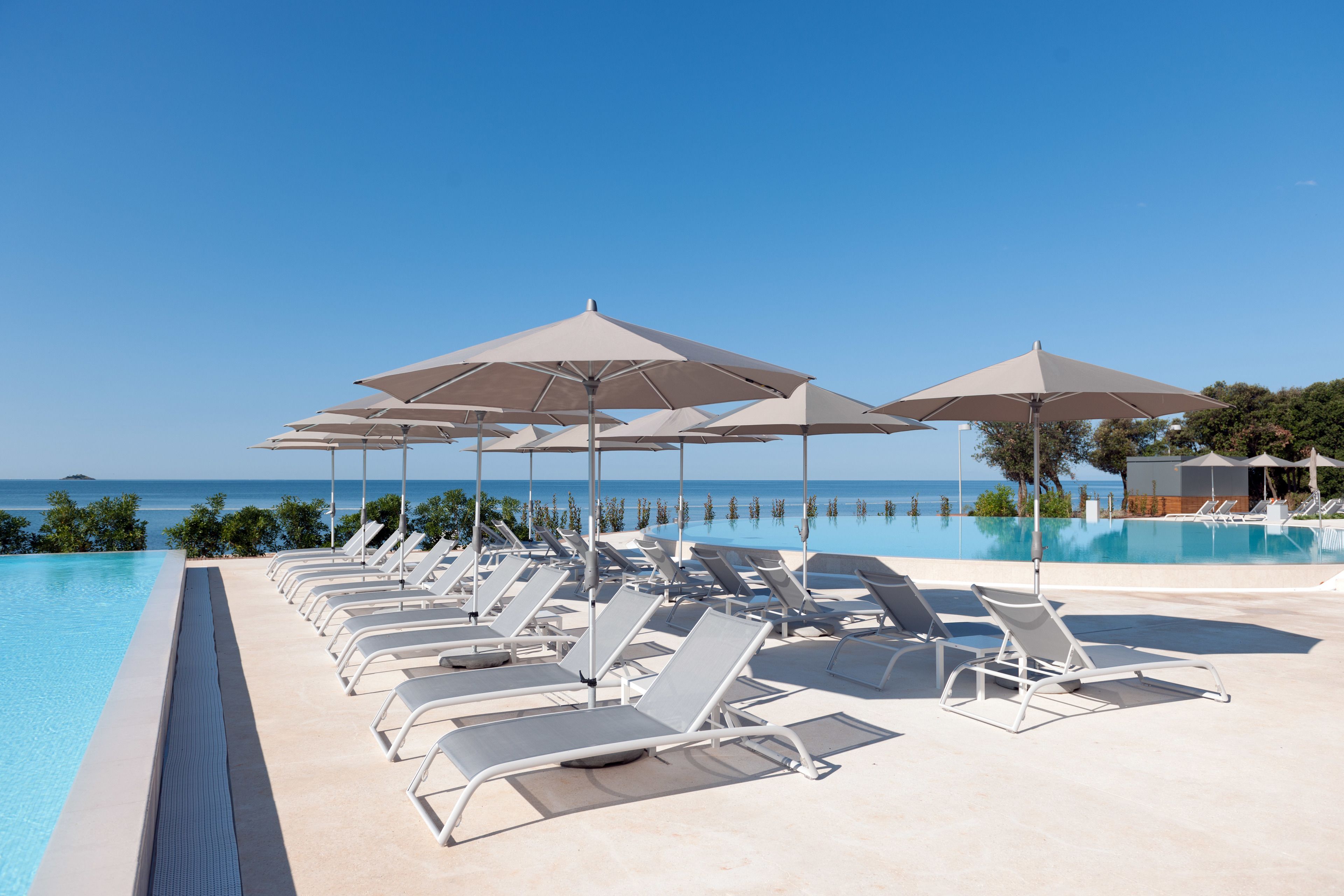 A poolside area with lounge chairs and umbrellas, a clear sky, and a view of the ocean.
