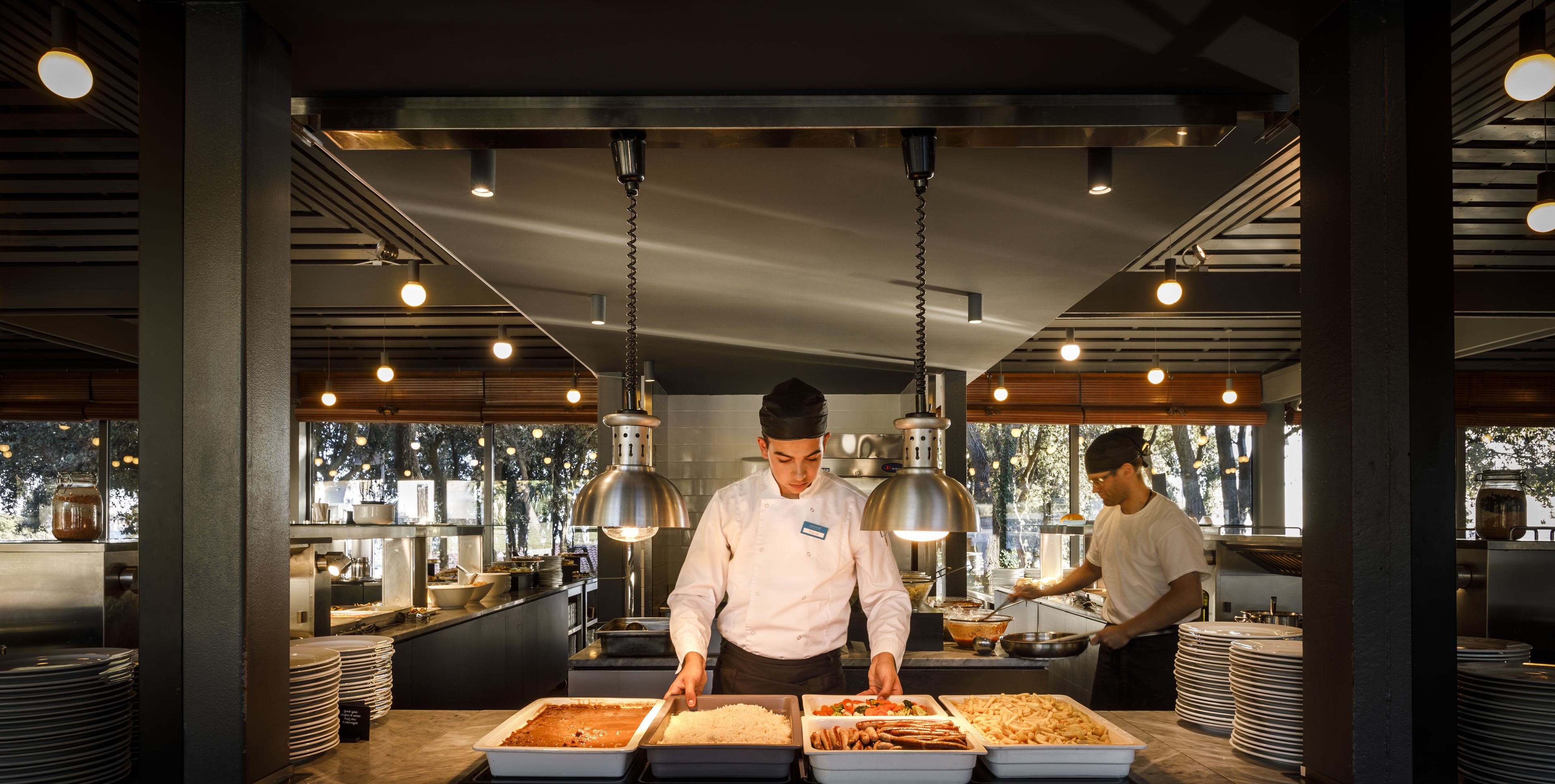 A modern kitchen with chefs preparing food, stainless steel appliances, and a view of the outdoors through large windows.